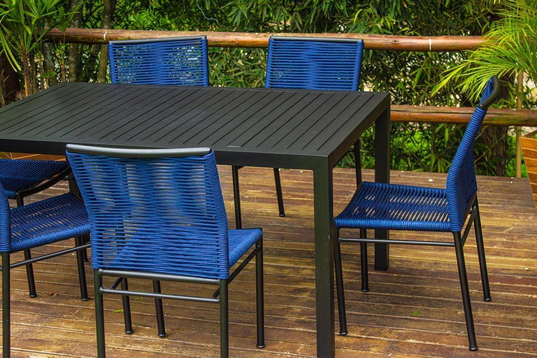 Mesa com cadeiras de trama azul em deck de madeira, em meio ao verde